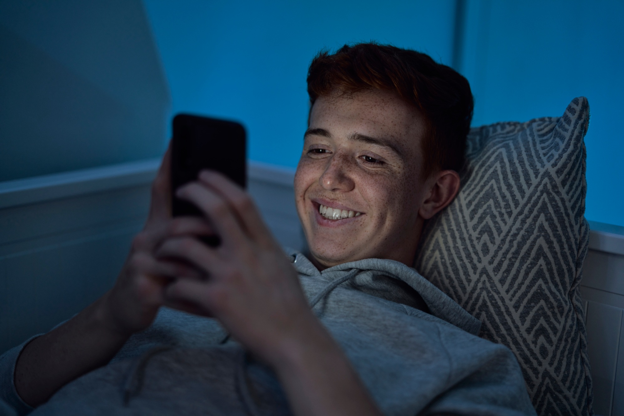 Cheerful caucasian teenage boy using mobile phone while lying in bed at night in his room
