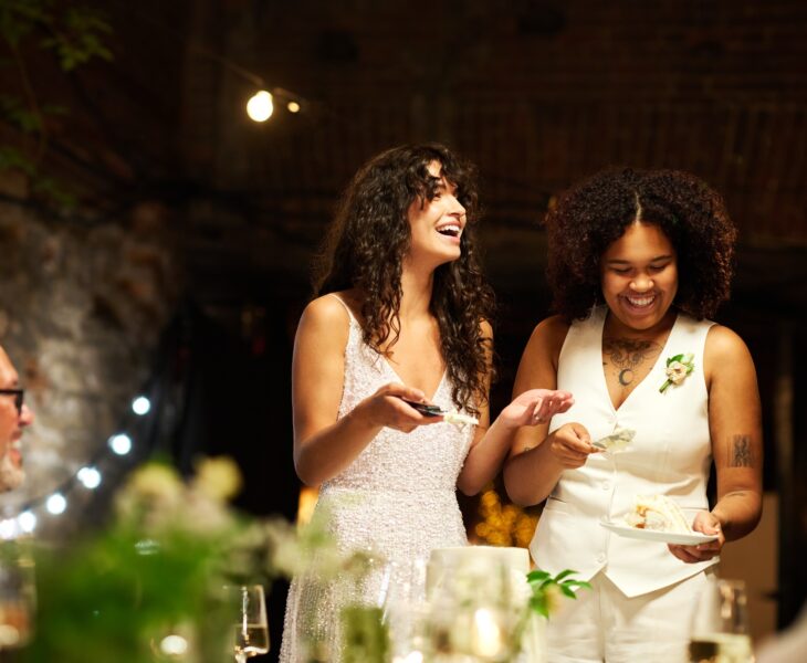 Cheerful young lesbian brides in wedding attire spreading cake among guests