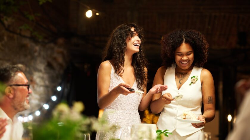 Cheerful young lesbian brides in wedding attire spreading cake among guests