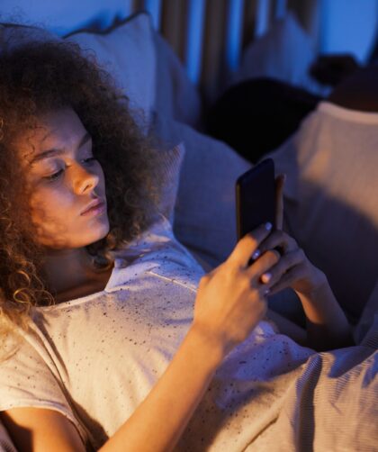 Young Woman Using Smartphone in Bed with Sleeping Husband