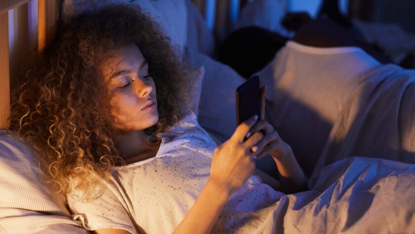 Young Woman Using Smartphone in Bed with Sleeping Husband