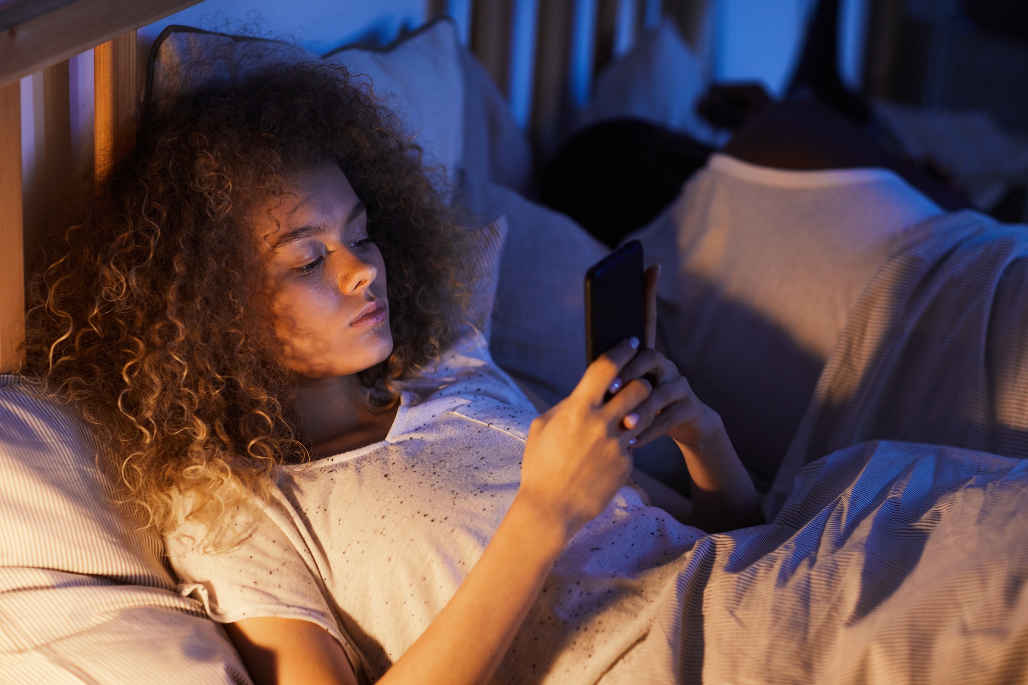 Young Woman Using Smartphone in Bed with Sleeping Husband