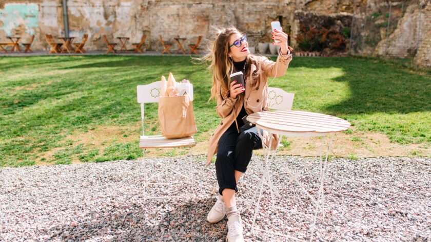 Attractive hipster girl wearing beige coat makes selfie for instagram profile during coffee break in